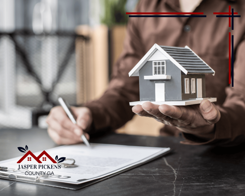 A person holds a model house while signing a document, symbolizing real estate transactions. The Jasper Pickens County GA logo emphasizes resources for understanding real estate commission settlements.